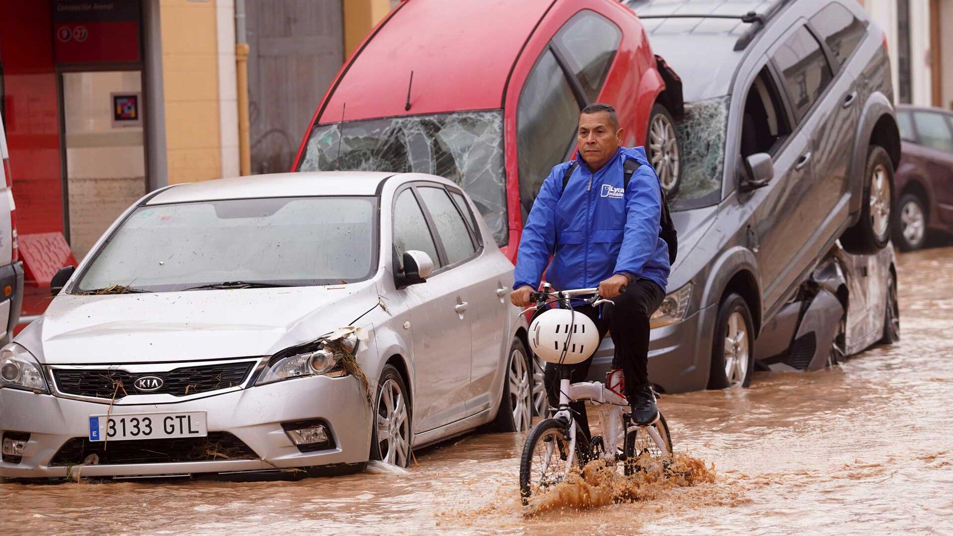 30.10.2024, Spanien, Valencia: Ein Mann radelt durch überflutete Straßen an Autos vorbei, die von den Wassermassen übereinander geschoben wurden