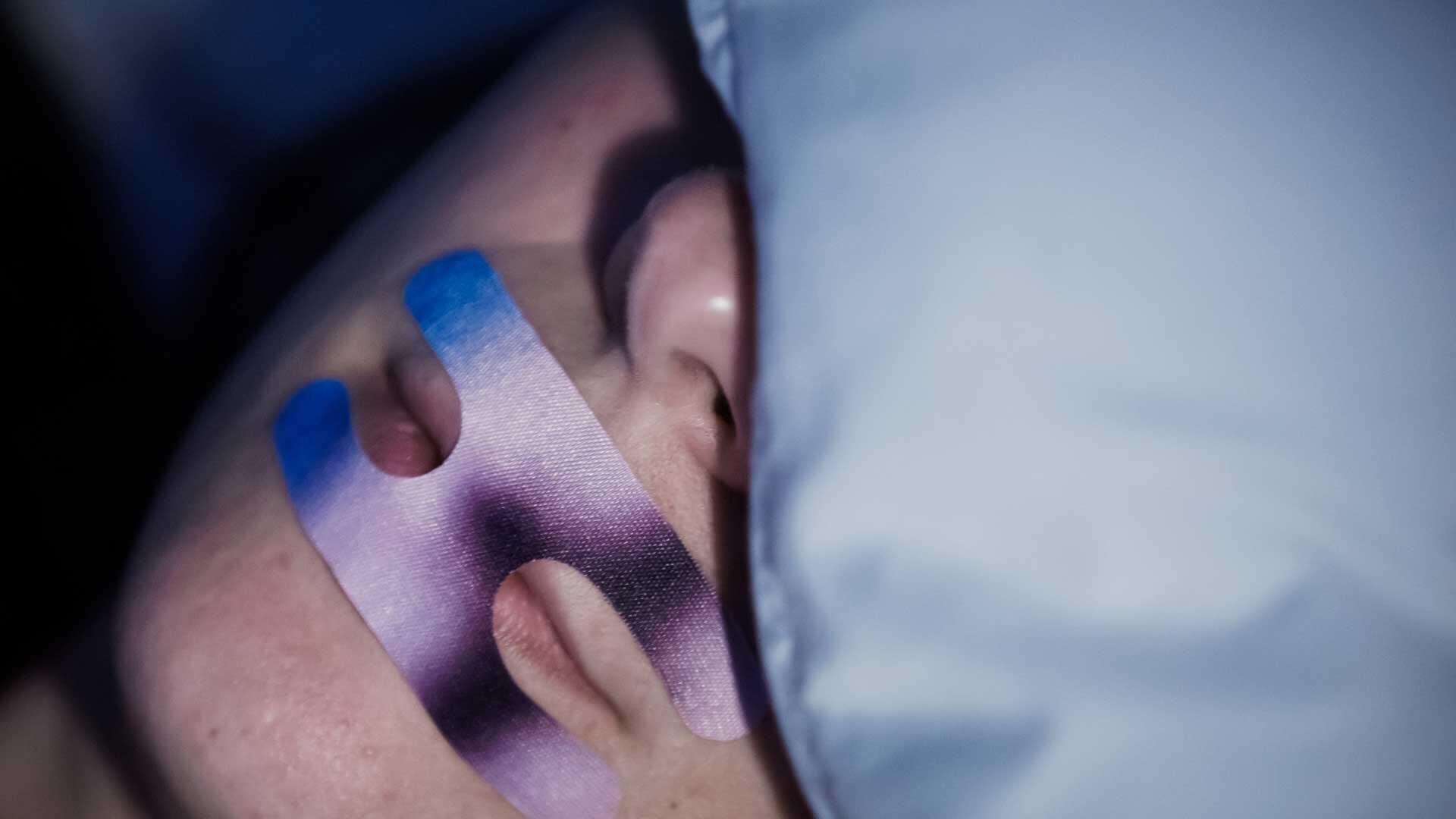 Eine Frau liegt mit einem Mundpflaster im Bett. Beim sogenannten Mouth Taping sollen ein Pflaster, Klebeband oder spezielle Tapes den Mund verschließen, so dass Menschen beim Schlafen statt durch den Mund durch die Nase atmen. 